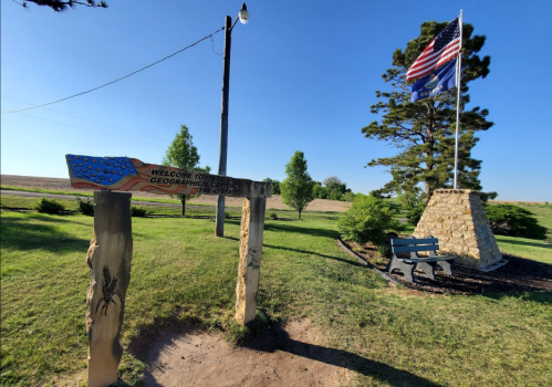 Sign welcoming visitors to the Geographic Center of the United States, with a flag and a bench nearby.