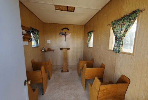 Interior of a small, simple chapel with wooden benches, a podium, and windows with patterned curtains.