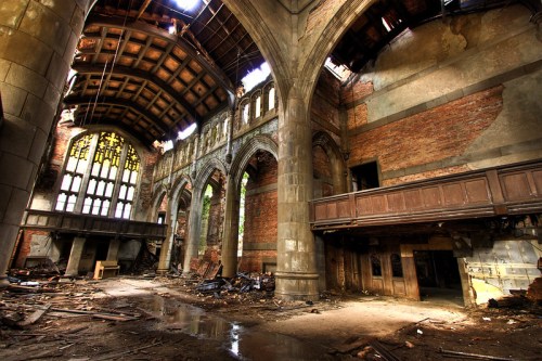 An abandoned, dilapidated interior of a building with high arches, broken windows, and debris scattered on the floor.