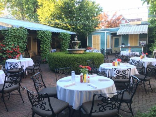 Outdoor dining area with tables, chairs, and a fountain, surrounded by greenery and colorful flowers.