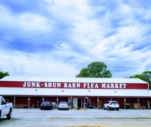 Exterior of Junk-Shun Barn Flea Market with a cloudy sky, featuring parked cars and a red barn-style building.