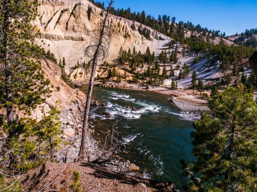 A serene river flows through a rocky landscape, surrounded by trees and steep cliffs under a clear blue sky.