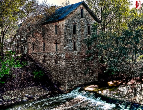 Abandoned stone building beside a flowing stream, surrounded by trees and greenery.