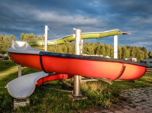 A red boat and a green water slide are mounted on poles in a grassy area under a cloudy sky.