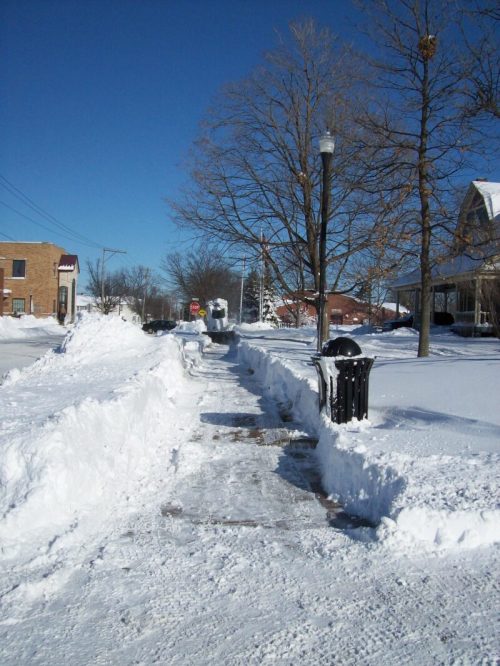 A snowy sidewalk lined with tall snowbanks, a clear blue sky, and a few trees in the background.
