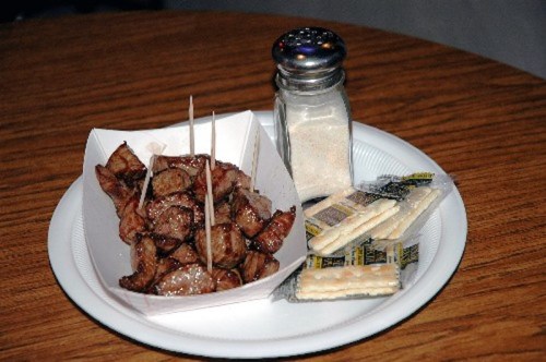 A plate with grilled meat cubes, salt shaker, and crackers on a wooden table.