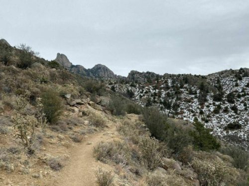A winding dirt path through rocky terrain, surrounded by sparse vegetation and distant mountains under a cloudy sky.