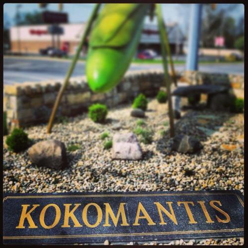 A sign reading "KOKOMANTIS" in front of a large green mantis sculpture in a landscaped area.