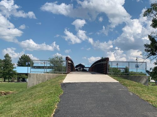 A paved path leads to a bridge over a grassy area, with blue skies and fluffy clouds in the background.