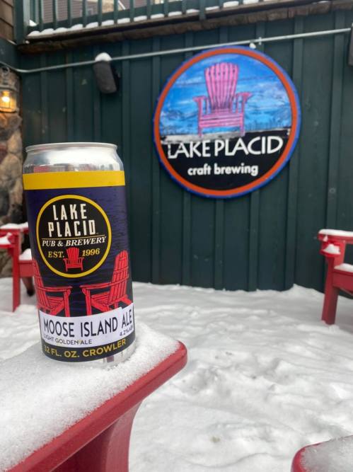 A can of Moose Island Ale in front of the Lake Placid craft brewing sign, with snow-covered red chairs nearby.