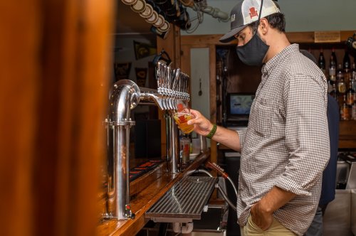 A man in a mask pours a drink from a tap at a bar, surrounded by bottles and bar decor.