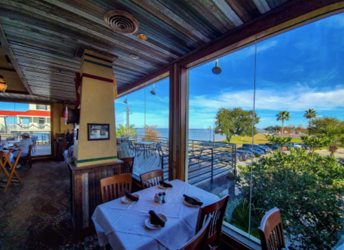 A restaurant interior with tables set for dining, large windows showcasing a sunny outdoor view with palm trees.