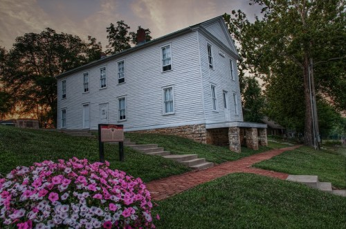 A historic white house on a grassy area, with a flower bed in front and a sign nearby, under a colorful sky.