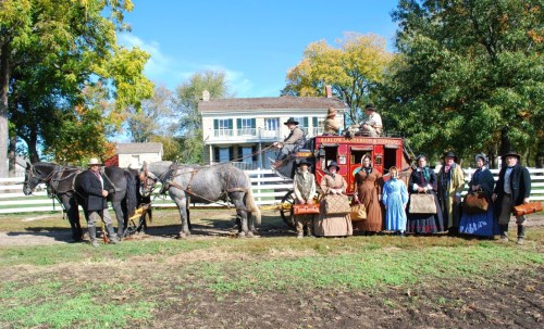 A group of people in historical costumes poses with a horse-drawn carriage in front of a vintage house and trees.