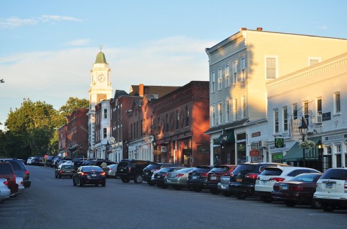 A charming street lined with parked cars and historic buildings, featuring a clock tower in the background.