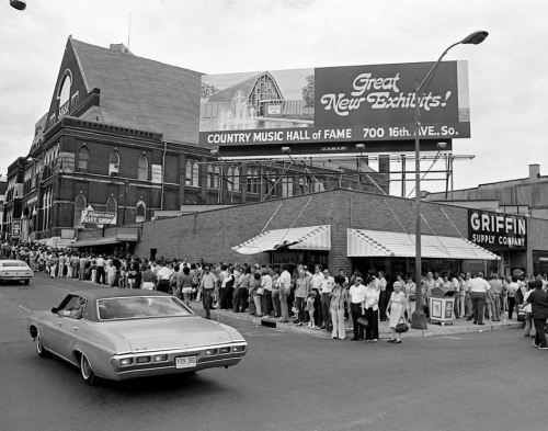 A black-and-white photo of a long line outside the Country Music Hall of Fame, with a vintage car passing by.