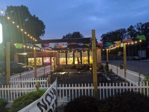 A cozy outdoor dining area with string lights and colorful banners, next to a restaurant at dusk.