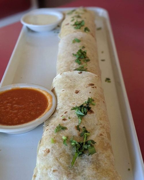 Two rolled flatbreads topped with chopped cilantro, served with a side of orange sauce and yogurt dip on a white plate.