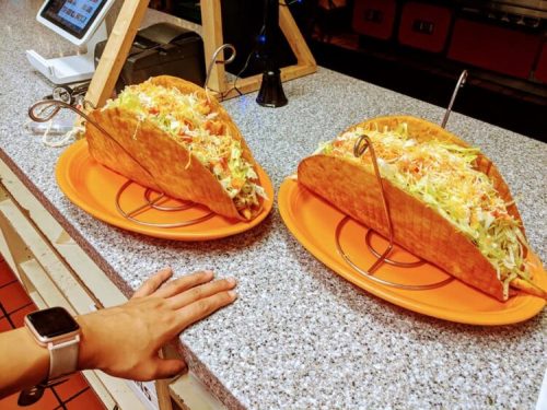 Two oversized tacos filled with lettuce, cheese, and tomatoes on orange plates, with a hand and watch in the foreground.
