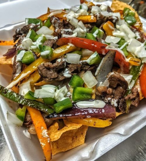 A plate of nachos topped with grilled beef, colorful bell peppers, onions, and drizzled with creamy sauce.