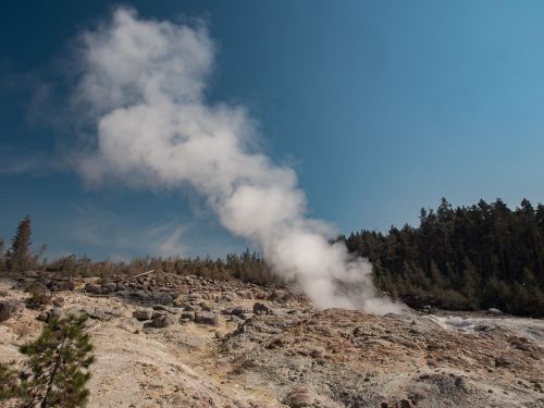 Steam rises from a geothermal area, surrounded by rocky terrain and trees under a clear blue sky.