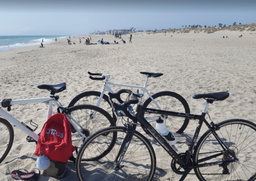 Two bicycles parked on a sandy beach with people in the background enjoying the sun and surf.