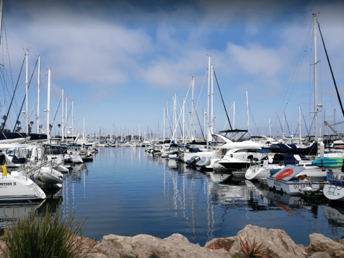 A serene marina filled with boats and yachts, reflecting in calm waters under a blue sky with scattered clouds.