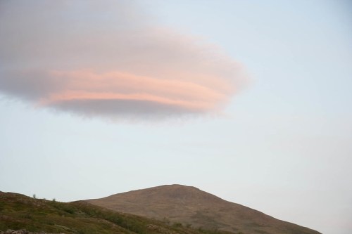 A soft, pink-tinged cloud hovers above a gentle, rolling hill under a clear sky.