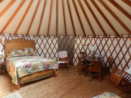 Cozy interior of a yurt featuring a bed, dining table, and wooden chairs with a warm, rustic design.