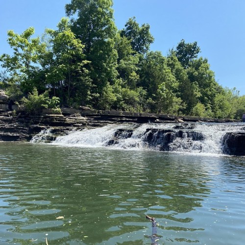 A serene waterfall cascades over rocks into a calm green pool, surrounded by lush trees under a clear blue sky.