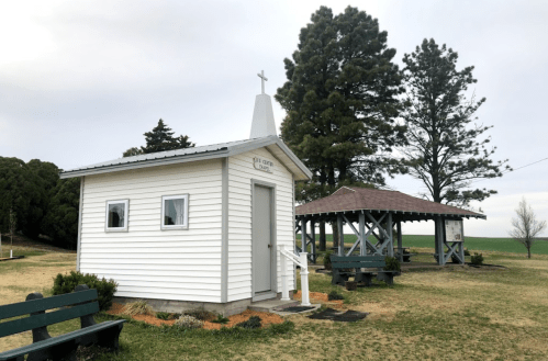 A small white chapel with a cross on top, surrounded by trees and a gazebo in a grassy area.