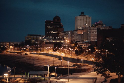 City skyline at dusk, featuring illuminated buildings and a waterfront path lined with lights.