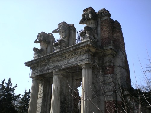 Ruins of a classical-style structure with three statues on top, partially covered in vines against a clear blue sky.