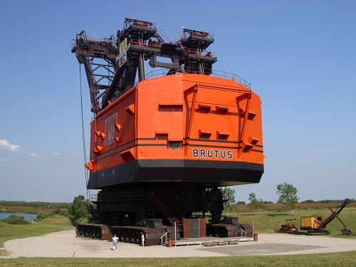 A large orange bucket-wheel excavator named Brutus, standing on tracks in an open area with blue sky and greenery.
