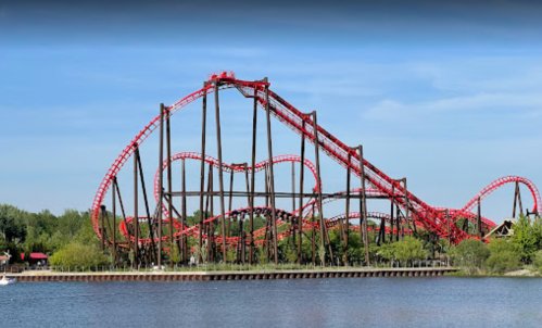 A red roller coaster with a heart-shaped loop towers over a lake, surrounded by trees and a clear blue sky.