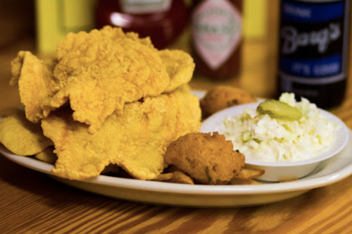 A plate of fried fish with hushpuppies, coleslaw, and condiments in the background.