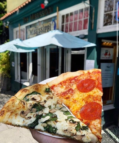 A hand holds two slices of pizza—one with pepperoni and the other with spinach and mushrooms—outside a restaurant.