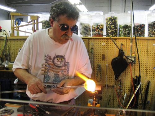 A man wearing sunglasses shapes molten glass in a workshop, with colorful beads and tools in the background.