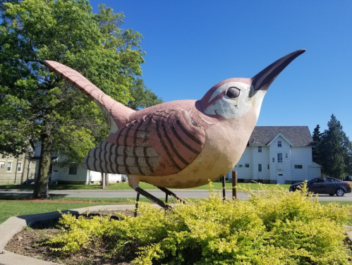 A large, colorful bird sculpture stands in a grassy area with trees and houses in the background under a clear blue sky.