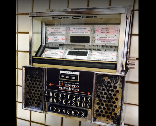 A vintage jukebox with a glass front, buttons for song selection, and a retro design, mounted on a tiled wall.