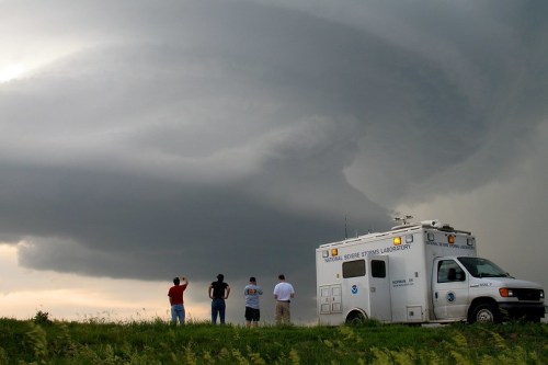 A group of people watches a dramatic storm cloud formation near a weather research vehicle.