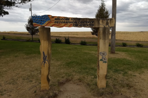 Sign reading "Welcome to the Geographical Center of the 48 States" with decorative elements, set in a grassy area.