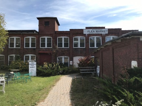 Exterior view of a brick building with a sign for a flea market, surrounded by greenery and outdoor seating.