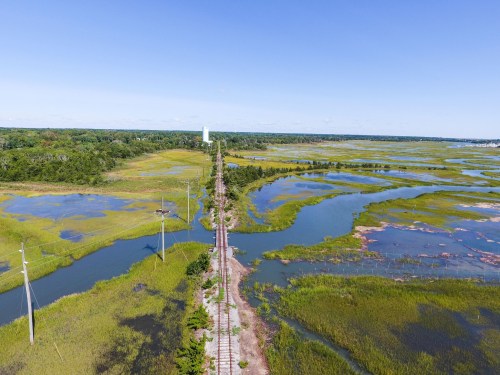 Aerial view of a railway track running through lush green marshlands and water bodies under a clear blue sky.