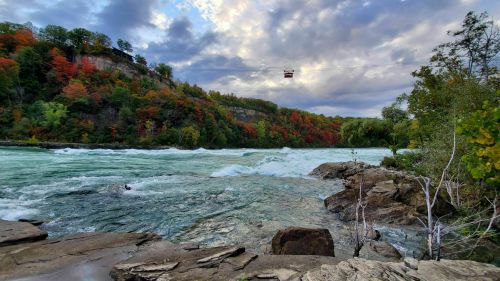 A scenic view of a river with rocky banks, surrounded by colorful autumn trees and a cloudy sky. A cable car is visible above.