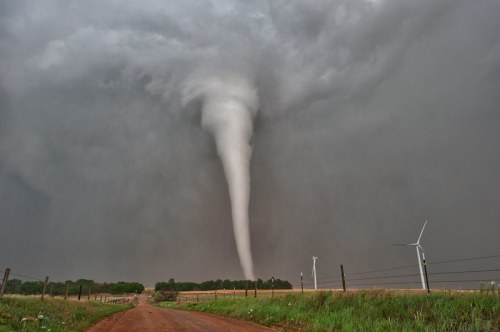 A tornado descends from dark storm clouds over a dirt road, with wind turbines visible in the background.