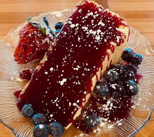 A slice of cheesecake topped with berry sauce, garnished with blueberries and a strawberry on a decorative plate.