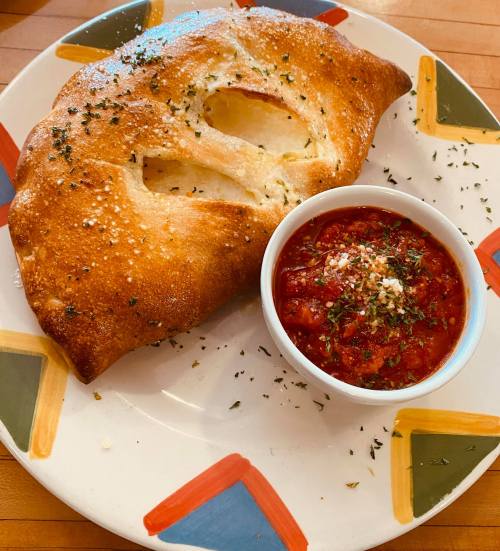 A golden-brown calzone on a colorful plate, served with a small bowl of marinara sauce sprinkled with herbs.