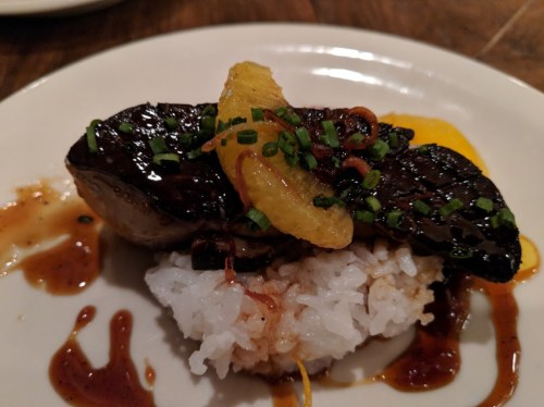 A plate featuring a piece of glazed meat atop white rice, garnished with orange slices and green onions.