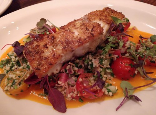 A beautifully plated dish featuring a piece of fish on a bed of colorful couscous salad with cherry tomatoes and microgreens.